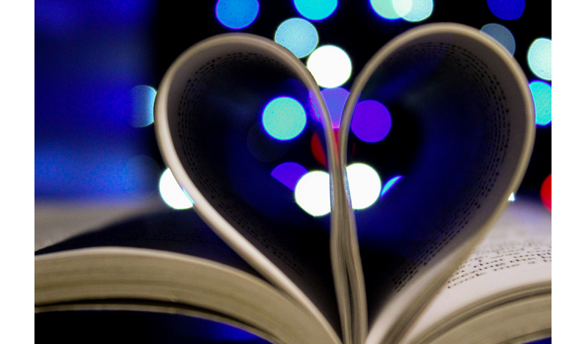 Aufgeschlagenes Buch mit eingeklappten Seiten in Herzform, im Hintergrund bunte Lichtpunkte – Symbol für die Liebe zum gedruckten Wort und handwerkliche Buchverarbeitung.
