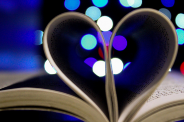 Aufgeschlagenes Buch mit eingeklappten Seiten in Herzform, im Hintergrund bunte Lichtpunkte – Symbol für die Liebe zum gedruckten Wort und handwerkliche Buchverarbeitung.