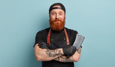 Mann mit rotem Vollbart, Schürze und Fleischermesser in der Hand posiert selbstbewusst vor hellem Hintergrund – Symbolbild für modernes Handwerk im Fleischereibereich.