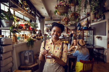 Junge Servicekraft steht freundlich lächelnd mit Notizblock in einem dekorativen Café – Symbolbild für Kundenkontakt, Gastfreundschaft und die vielfältigen Lehrberufe im österreichischen Tourismus.